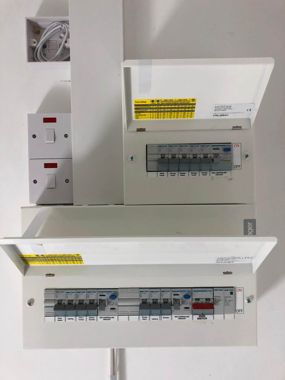 A close-up of an open consumer unit (fuse box) showing circuit breakers and voltage information mounted on a white wall.