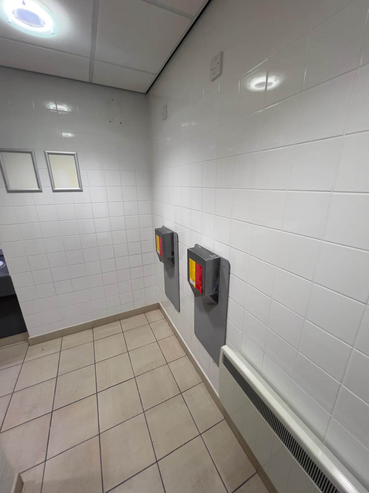View of a tiled bathroom corner featuring a grey hand dryers mounted on white textured walls.